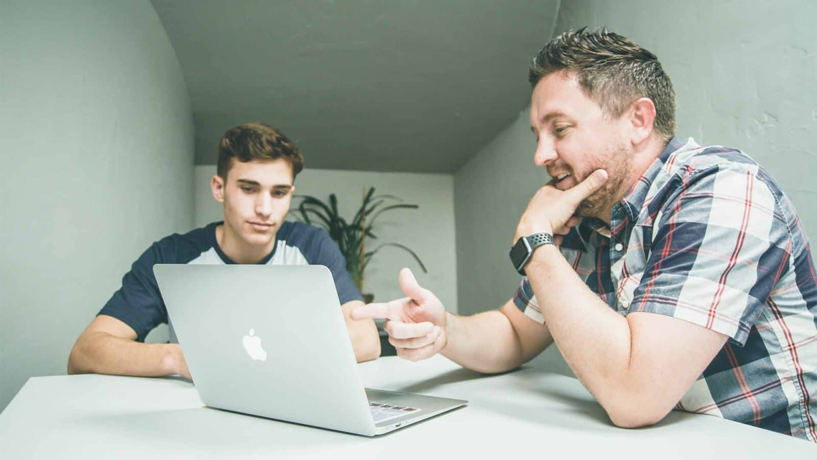 Una persona è seduta in casa utilizzando un laptop e un computer, il volto umano illuminato dallo schermo.