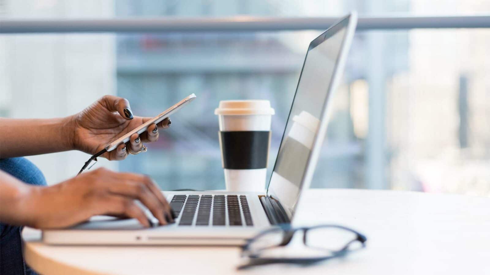 A pessoa está usando um laptop em uma mesa enquanto toma uma xícara de café.