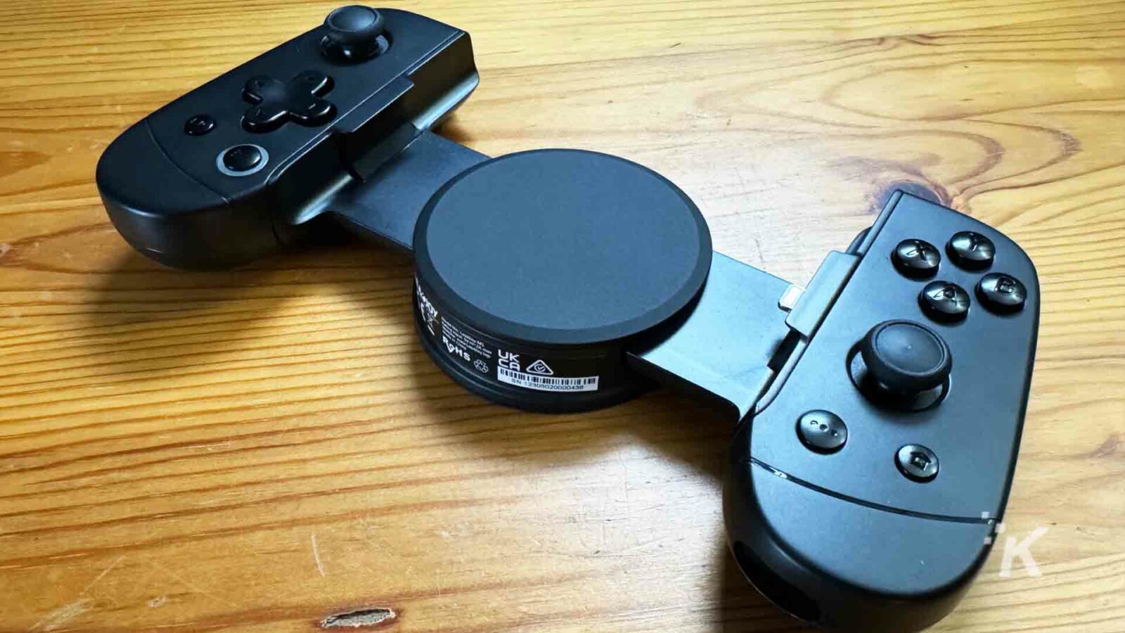 Un controller di gioco con joystick, dispositivo elettronico e telecomando è posizionato su un tavolo di legno all'interno.