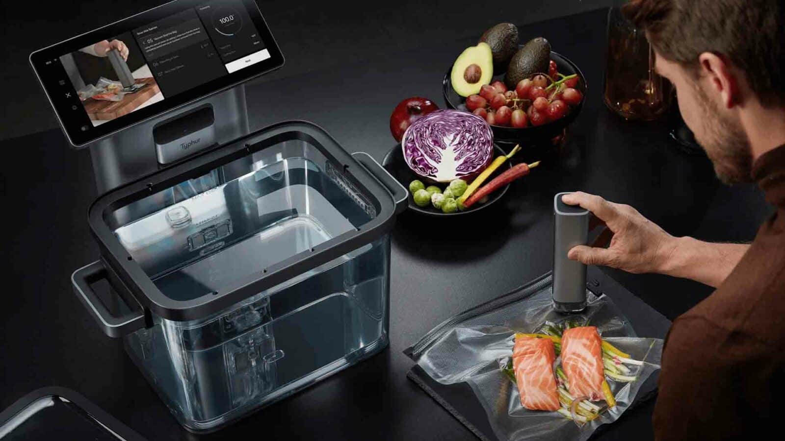 Seseorang sedang menyegel salmon dengan vakum di stasiun typhur sous-vide. Kompor sous-vide hadir bersama sayuran segar, alpukat, dan anggur di meja dapur modern.