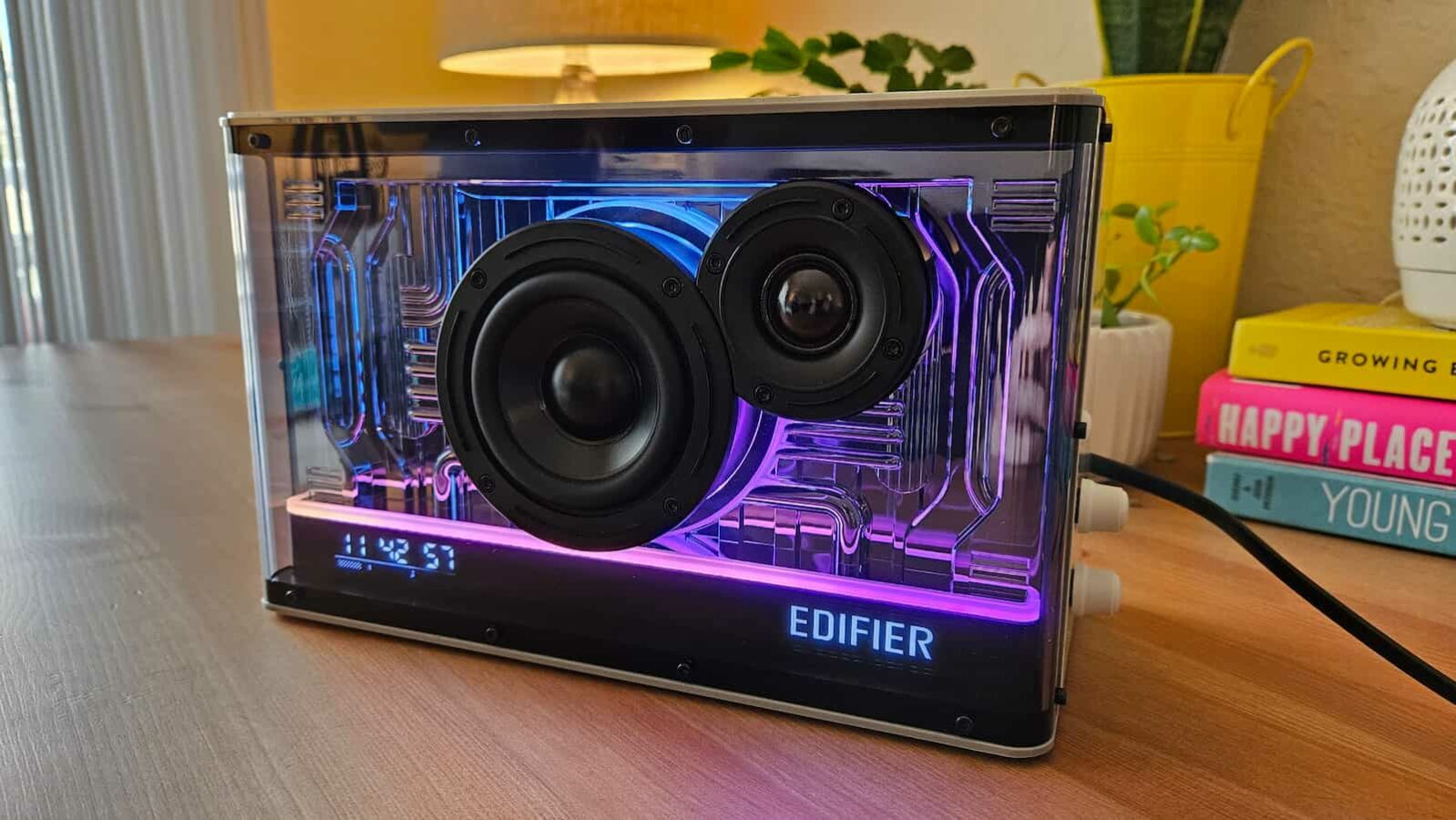 Un altavoz edificable transparente con partes internas iluminadas en violeta y azul se muestra sobre una superficie de madera, con libros y decoración al fondo.