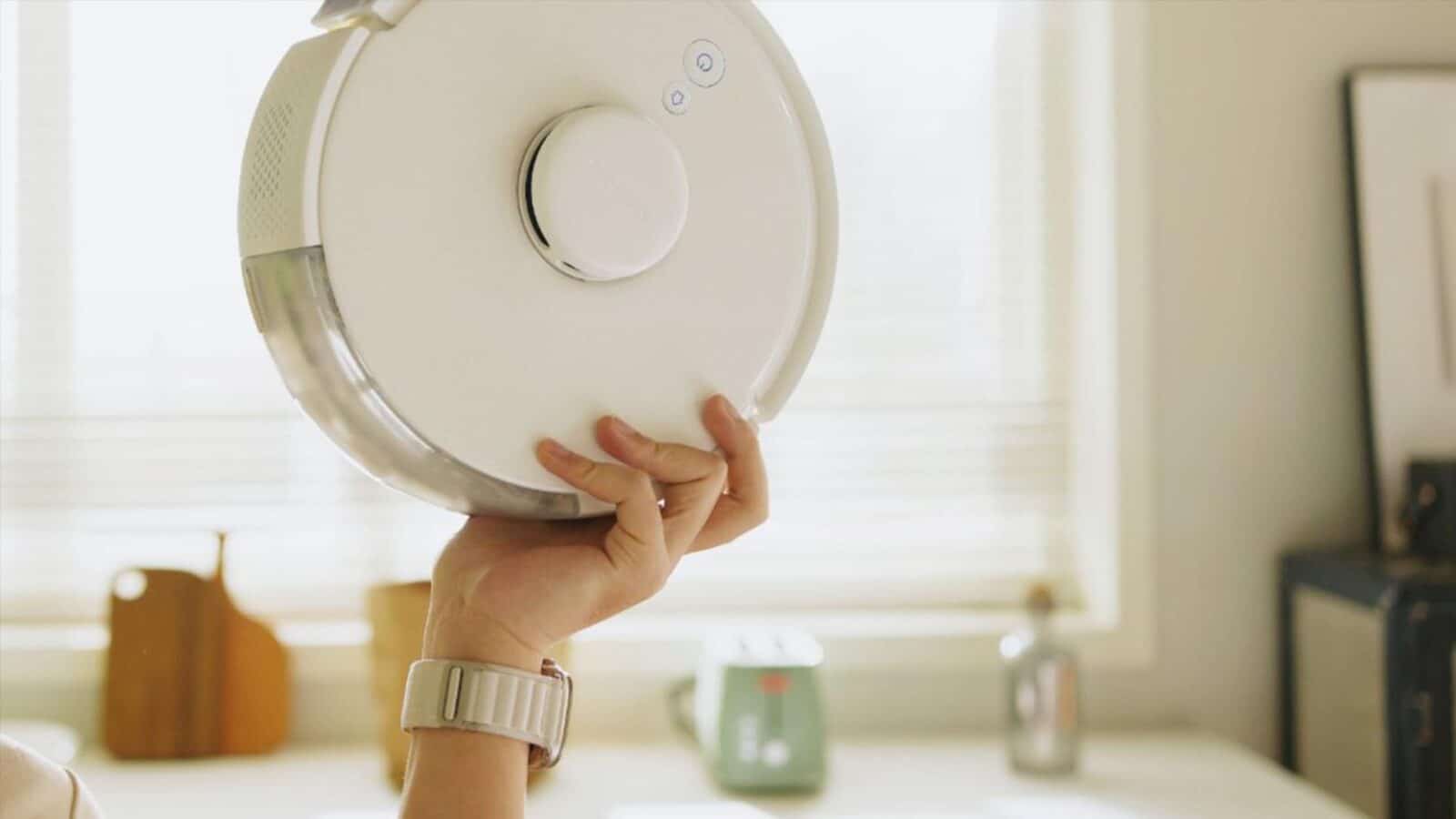 Une personne tient un robot aspirateur blanc dans une pièce bien éclairée avec des ustensiles de cuisine en arrière-plan. Il semble moderne et propre.