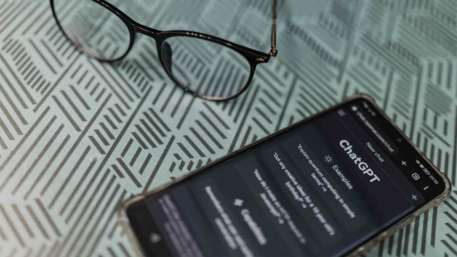 chatgpt アプリを開いたスマートフォンがメガネの隣の模様のある面に置かれており、仕事や勉強のシナリオを示唆しています。