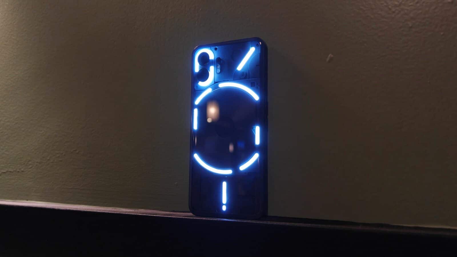 Un teléfono inteligente con un diseño futurista iluminado en su parte posterior descansa sobre una superficie oscura contra una pared con un débil reflejo de luz.