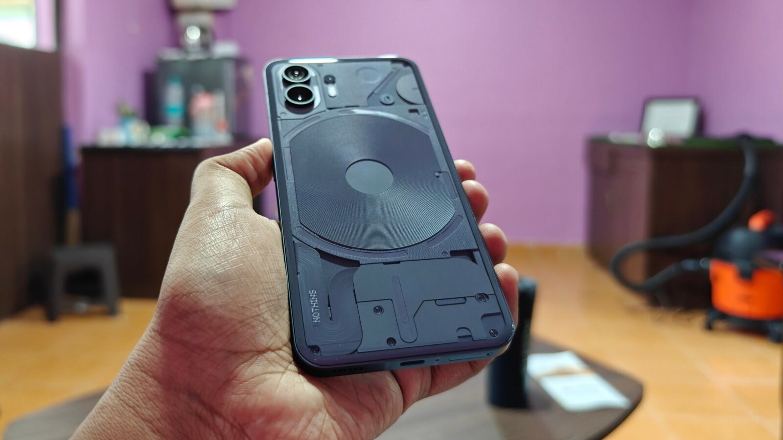 La mano de una persona sostiene un teléfono inteligente con una funda transparente que revela detalles internos. El fondo es una habitación borrosa con paredes y muebles de color violeta.