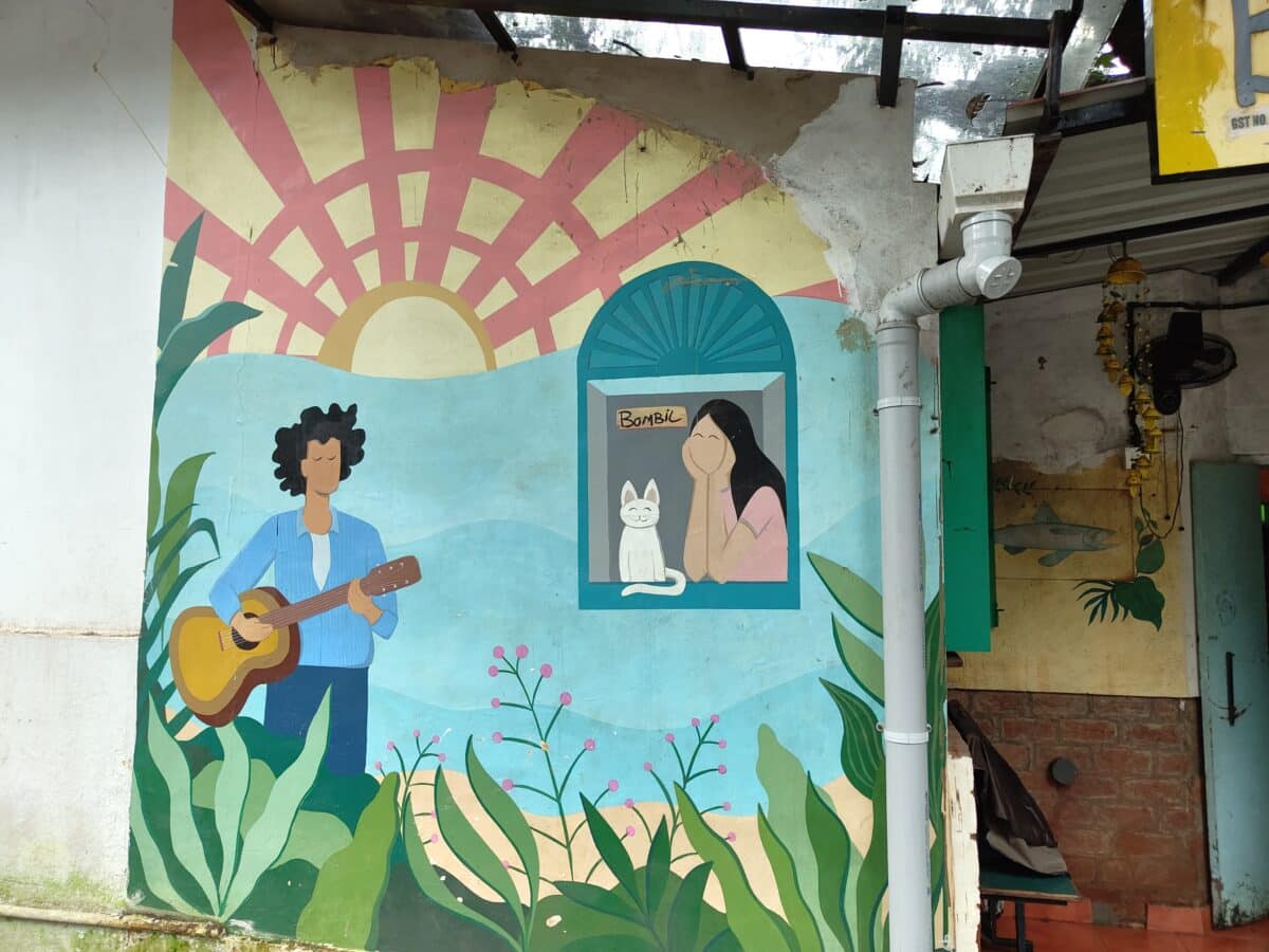 L'immagine mostra un murale colorato con una persona che suona la chitarra, un'immagine incorniciata con un'altra persona e un gatto, circondati da motivi vegetali.