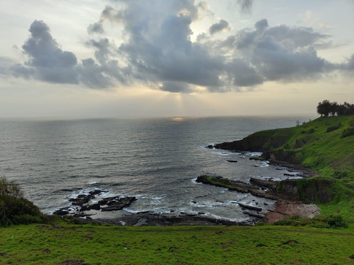 Esta imagem mostra uma paisagem costeira serena com falésias verdejantes, formações rochosas em camadas na costa e um raio de sol atravessando as nuvens sobre o mar.