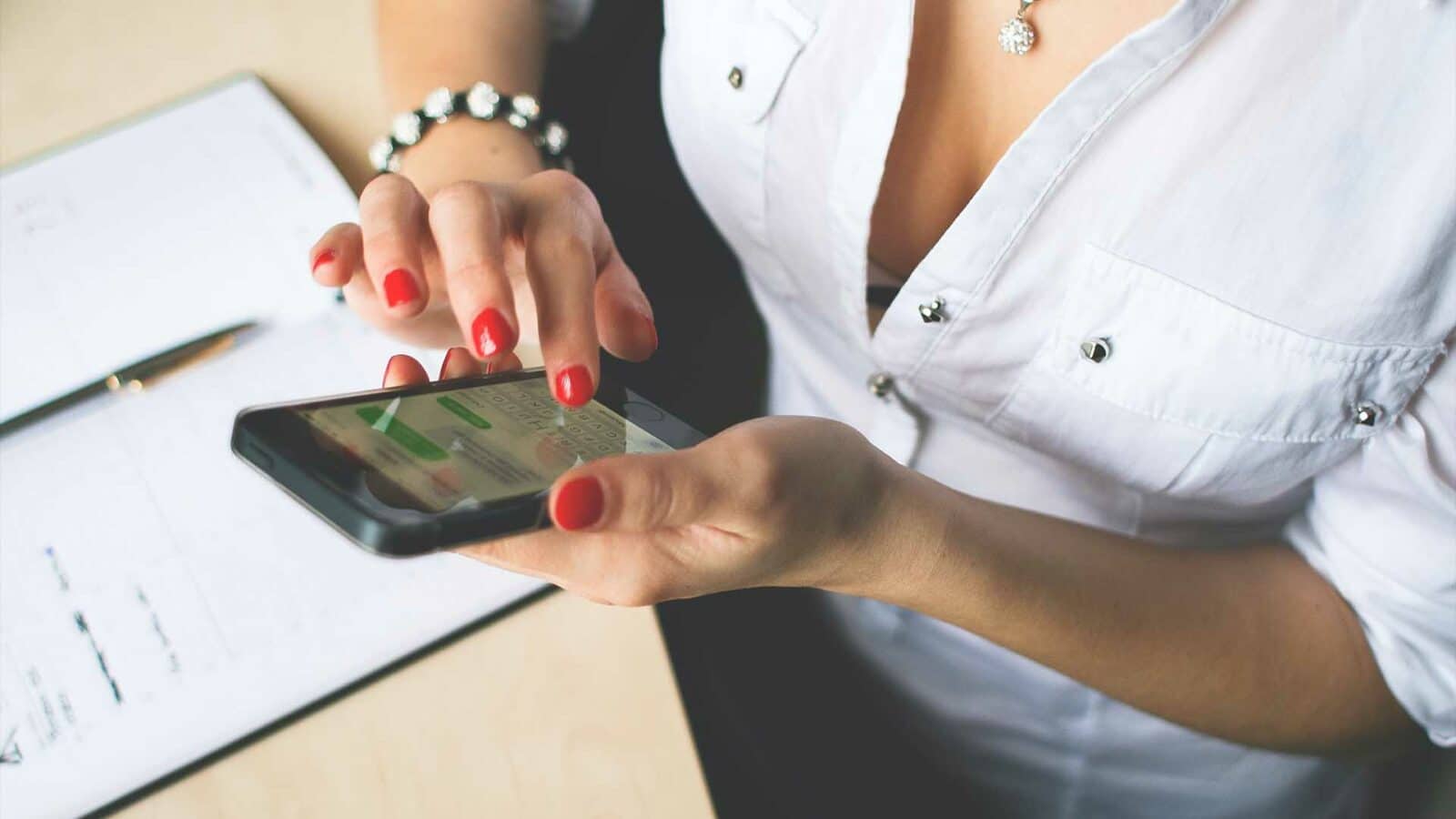 Una persona con lo smalto rosso usa uno smartphone su una scrivania con documenti e penna. Il focus è sulle mani e sul telefono.