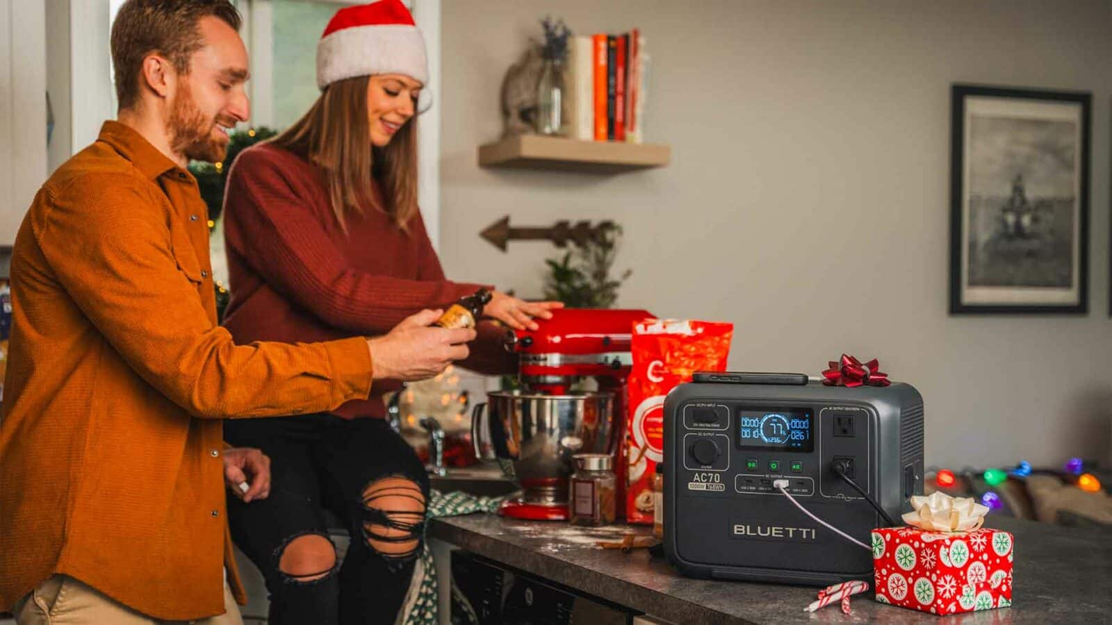 Dos personas disfrutando de la repostería navideña junto con una batidora. Una central eléctrica portátil y regalos envueltos para regalos sugieren un ambiente festivo con adornos navideños.