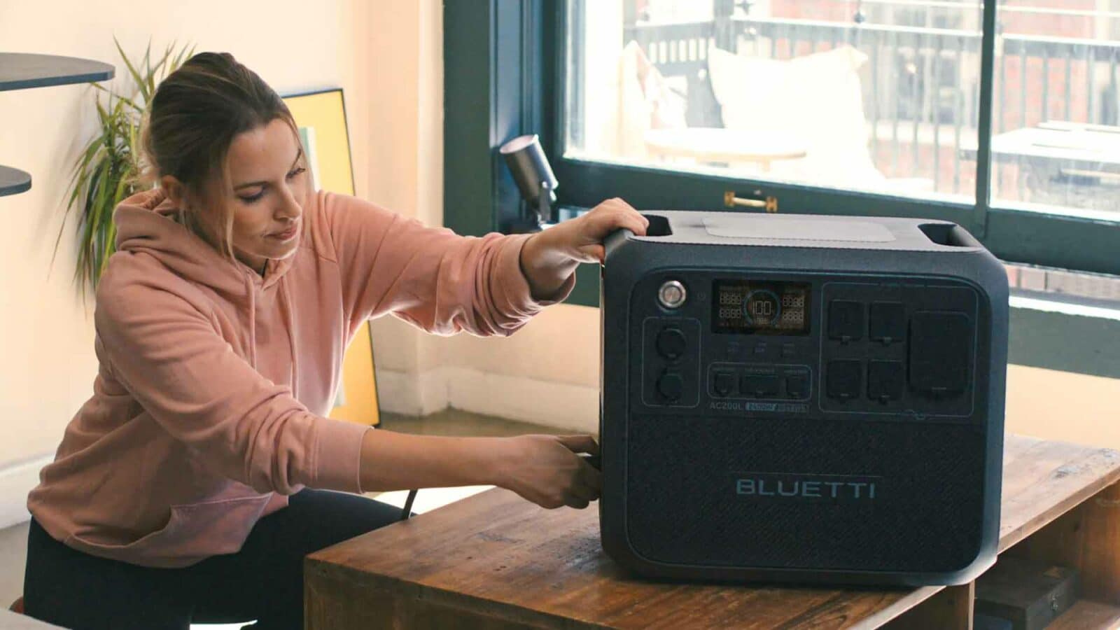 Una persona interactúa con una gran unidad de batería portátil con la etiqueta "bluetti" sobre una mesa de madera en el interior, cerca de una ventana soleada con persianas. La central eléctrica de 200 litros.