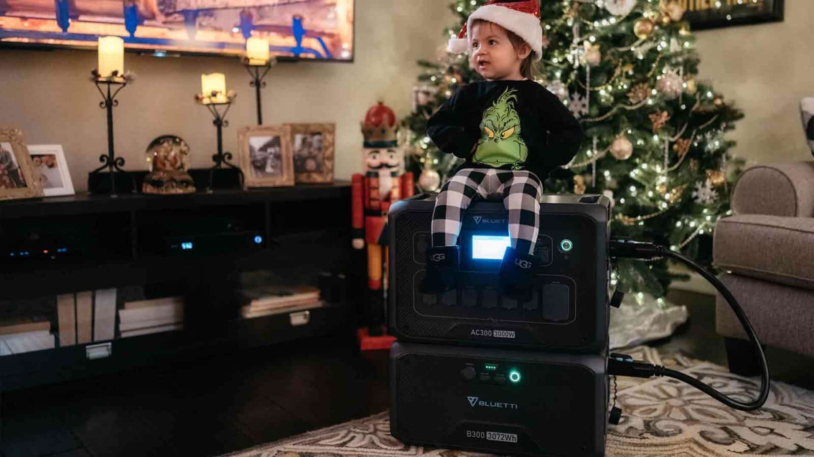 Un niño con un gorro de Papá Noel y un suéter de Grinch está sentado encima de una central eléctrica portátil en una habitación acogedora y decorada con un árbol de Navidad.