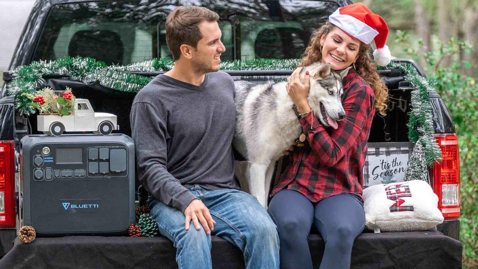 Dos adultos y un perro están sentados en la caja de una camioneta adornada con adornos festivos, que incluyen un gorro navideño, guirnaldas y una almohada navideña.