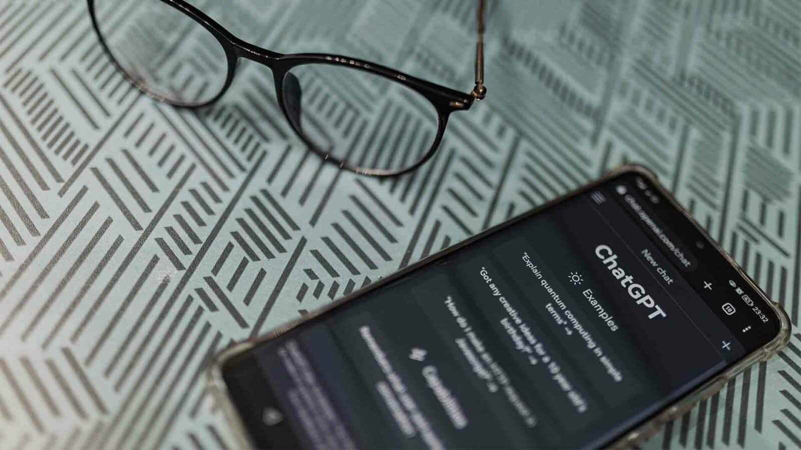 Ein Smartphone mit geöffneter Chatgpt-App liegt auf einer gemusterten Oberfläche neben einer Brille und deutet auf ein Arbeits- oder Lernszenario hin.