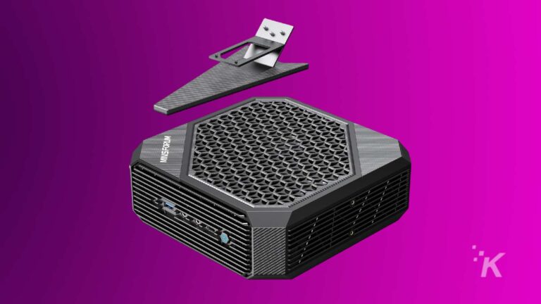 Um computador desktop compacto cinza grafite com design de ventilação hexagonal em um fundo roxo vívido, acompanhado por um suporte externo correspondente com um cartão branco flutuante com um rosto sorridente.