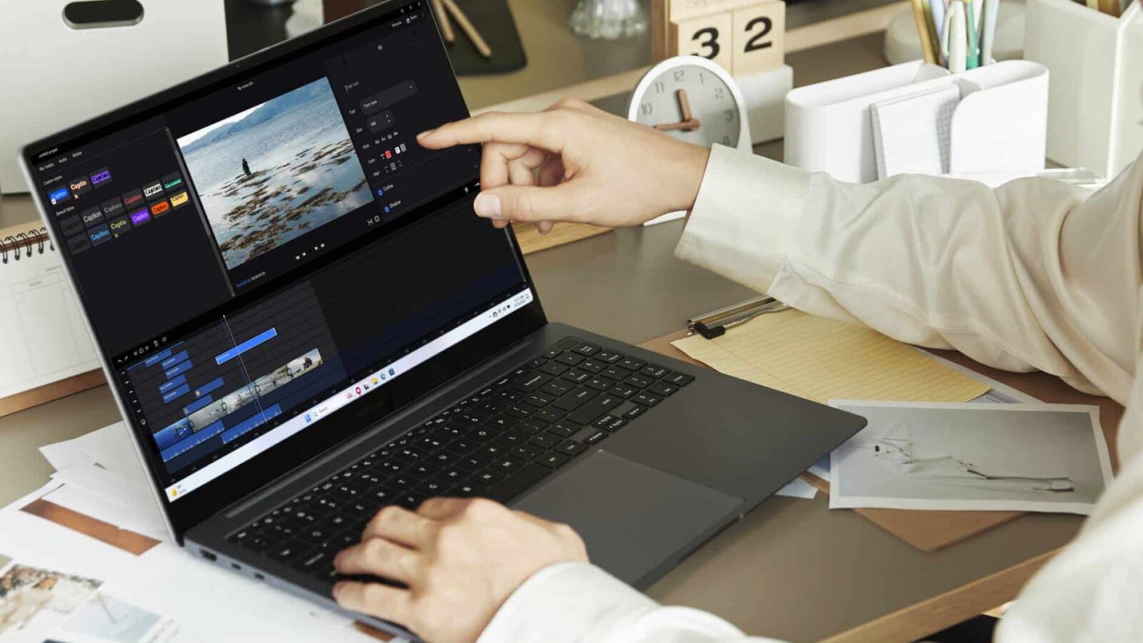 Una persona editando un vídeo en una computadora portátil con software de edición de video, apuntando a la pantalla, sobre un escritorio con material de oficina y fotografías.
