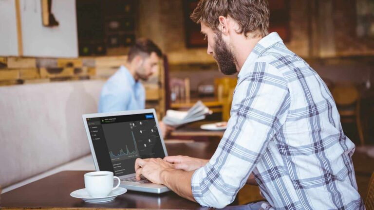 Um homem de camisa xadrez trabalhando em um laptop com gráficos analíticos na tela, sentado em uma mesa de madeira em um café com uma xícara de café, com outra pessoa trabalhando em segundo plano.