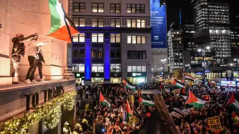 Massa berunjuk rasa dengan bendera Palestina di kota pada malam hari.